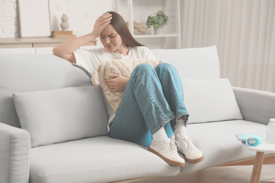 Vrouw die menstruatieklachten ervaart zoals hoofdpijn en buikkrampen en uitgeput op de bank zit.